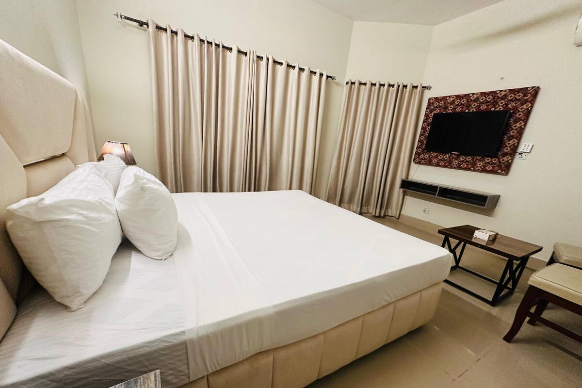 A hotel room with a large bed, a bedside table, a chair, a TV mounted on the wall, and curtains.
