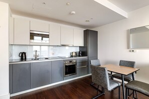 A modern kitchen with grey cabinets, a built-in oven, and a dining area with a table and chairs. - Blueground | South Quays, fitness center, nr Thames (London)