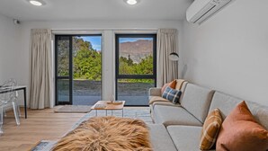 A modern living room with a sofa, a coffee table, and a dining area. There are large windows with a view of greenery and mountains. - Arawata Alpine Views - 2BDRM luxury (Queenstown)