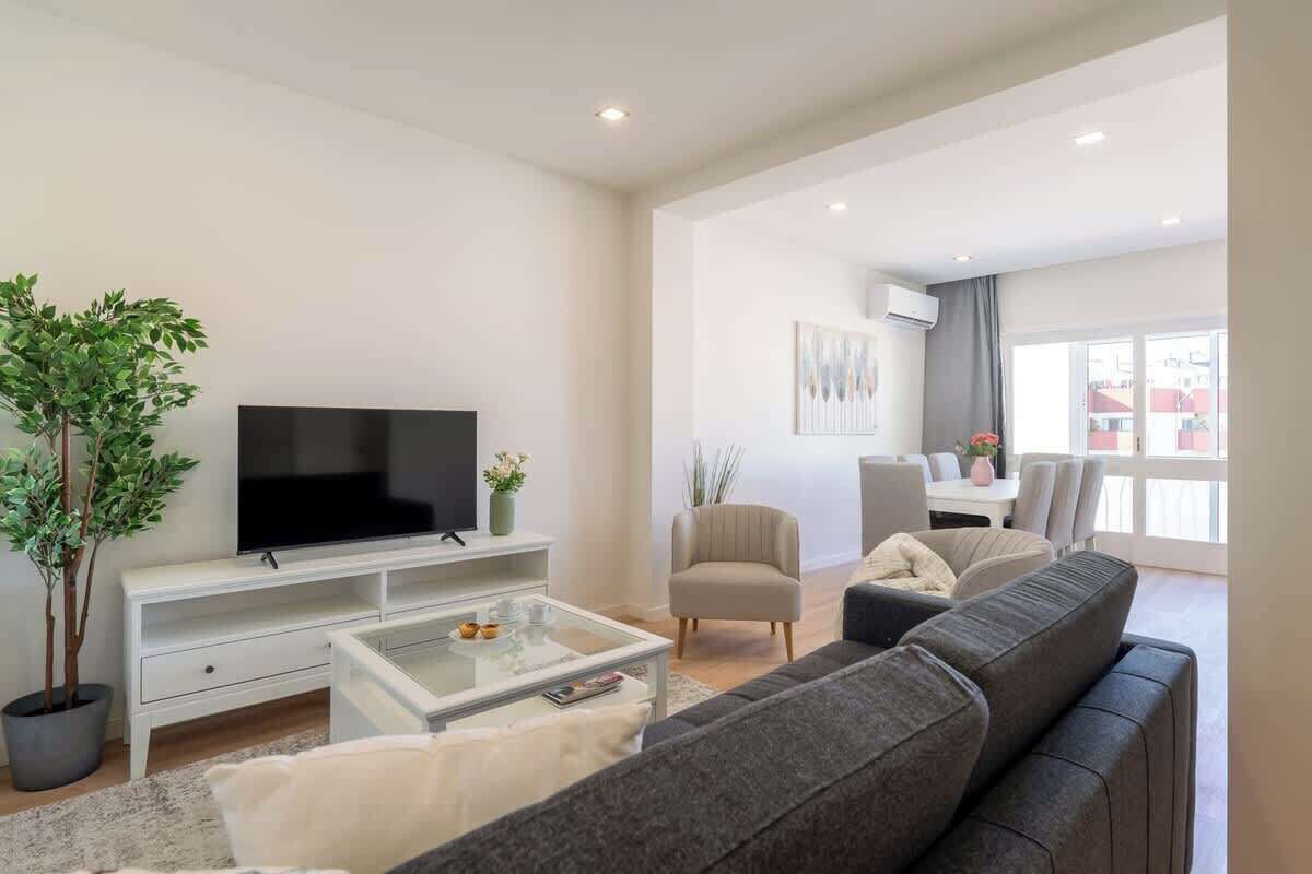 A modern living room with a flat-screen TV, a sofa, a coffee table, and a dining area with a table and chairs.