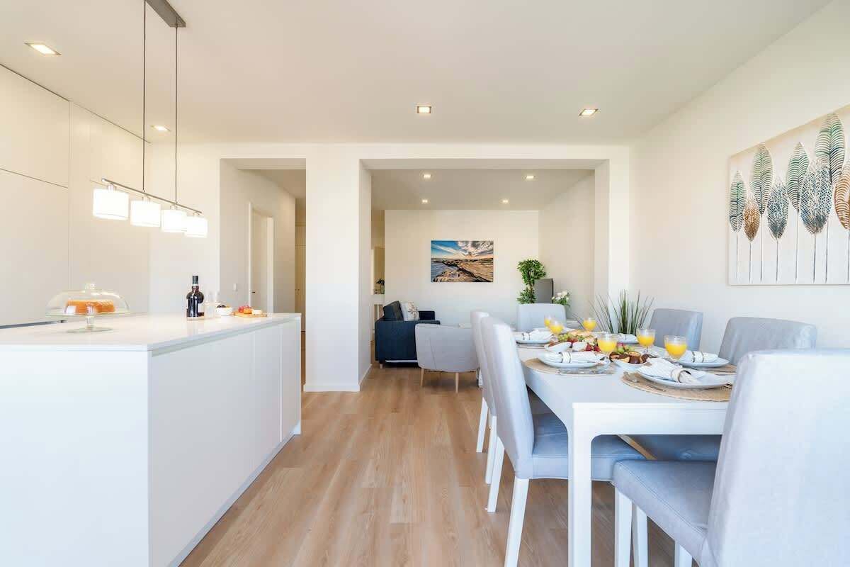 A modern kitchen with a central island, a dining area with a white table and chairs, and a living space with a sofa and a painting on the wall.