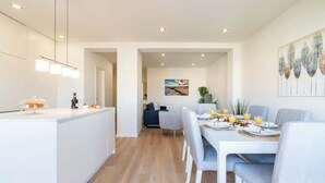 Une cuisine moderne avec un îlot central, un coin repas avec une table et des chaises blanches, et un espace de vie avec un canapé et un tableau au mur.