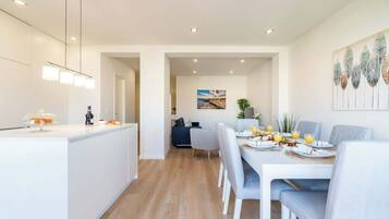A modern kitchen with a central island, a dining area with a white table and chairs, and a living space with a sofa and a painting on the wall.