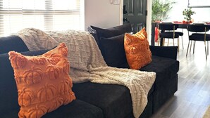 Ein Wohnzimmer mit einem dunkelgrauen Sofa, einer hellen Decke und zwei orangefarbenen Kissen. Im Hintergrund ist ein Essbereich mit Tisch und Stühlen zu sehen.