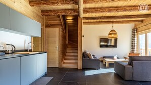 A modern kitchen with grey cabinets and a wooden staircase leading to a living area with a grey sofa and a TV. - MAKALU LODGE - BEAUFORT (BEAUFORT)