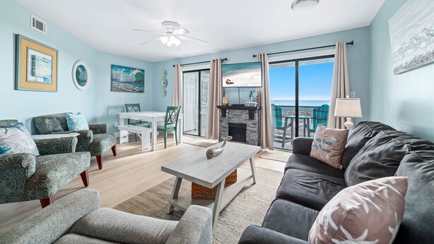 Een ruime woonkamer met een grijze bank, bijpassende fauteuils, een salontafel en een open haard. Op de achtergrond is een eethoek met een witte tafel en stoelen te zien.