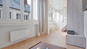 Amplio salón con grandes ventanales, un radiador, una mesa de centro de madera y un pequeño altavoz.