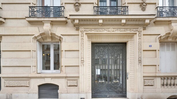 Fachada de edificio con una puerta grande y ornamentada, marcos de ventanas decorativos y un pequeño balcón con barandilla de hierro forjado.