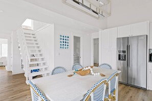 A modern kitchen with white cabinetry, a stainless steel refrigerator, and a dining area with a white table and blue and white checkered chairs. - Escape to Sandy Dreams, a beautifully remodeled villa perfectly situated on Bald Head Island. (Bald Head Island)