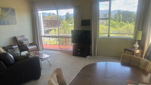 Una sala de estar con sofá, mesa de comedor, televisor y balcón con vistas a zonas verdes.