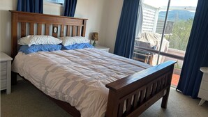 Un dormitorio con cama de madera, ropa de cama blanca, una mesita de noche y una ventana con cortinas que ofrece vistas a edificios y árboles.