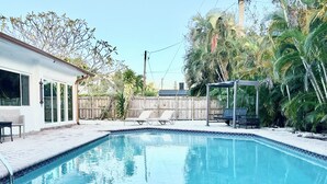 Una piscina con borde de azulejos, rodeada por un patio de ladrillo. Hay sillas de descanso y una zona de estar cubierta.
