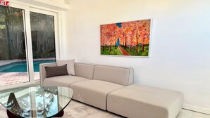 Un salón con un sofá beige, una mesa de centro de vidrio y un cuadro de árboles.