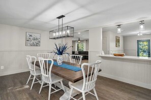 En matplats med ett träbord, vita stolar, en vas med blått lövverk och en hängande lampa.
