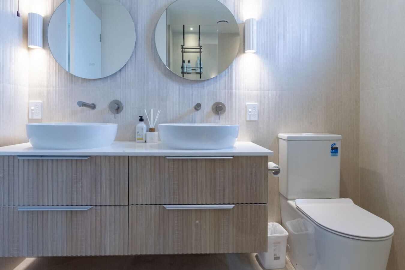 Une salle de bain moderne avec deux vasques sur pied, des toilettes et deux miroirs fixés au mur.