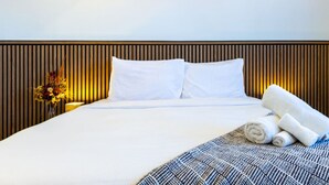 Une chambre d’hôtel avec un lit, du linge de lit blanc, une tête de lit en bois, un vase avec des fleurs et des serviettes enroulées.