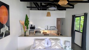 A modern bedroom with a bed, bedside table, and a view of a compact kitchen with a dining area. - Brisas del Mar Bocas (Bastimentos)