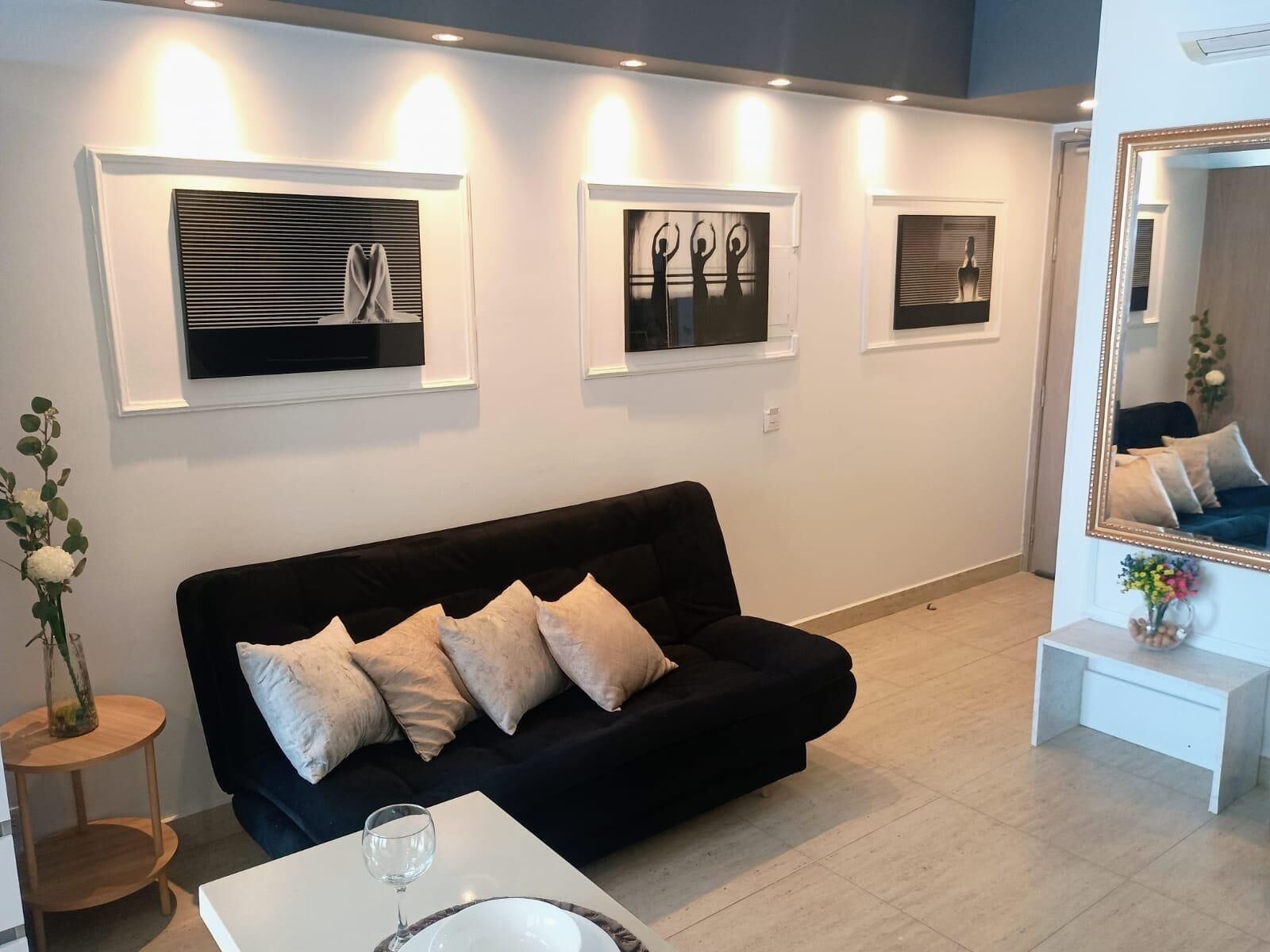 A modern living room with a black sofa, white cushions, a glass coffee table, and framed artwork on the wall.
