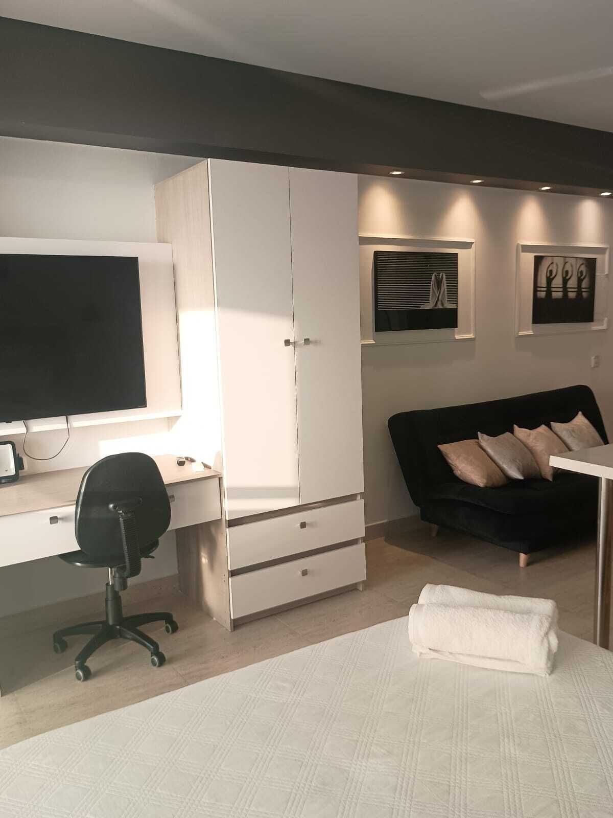 A modern bedroom with a bed, a desk with a computer, a black office chair, a black sofa, and a white wardrobe.