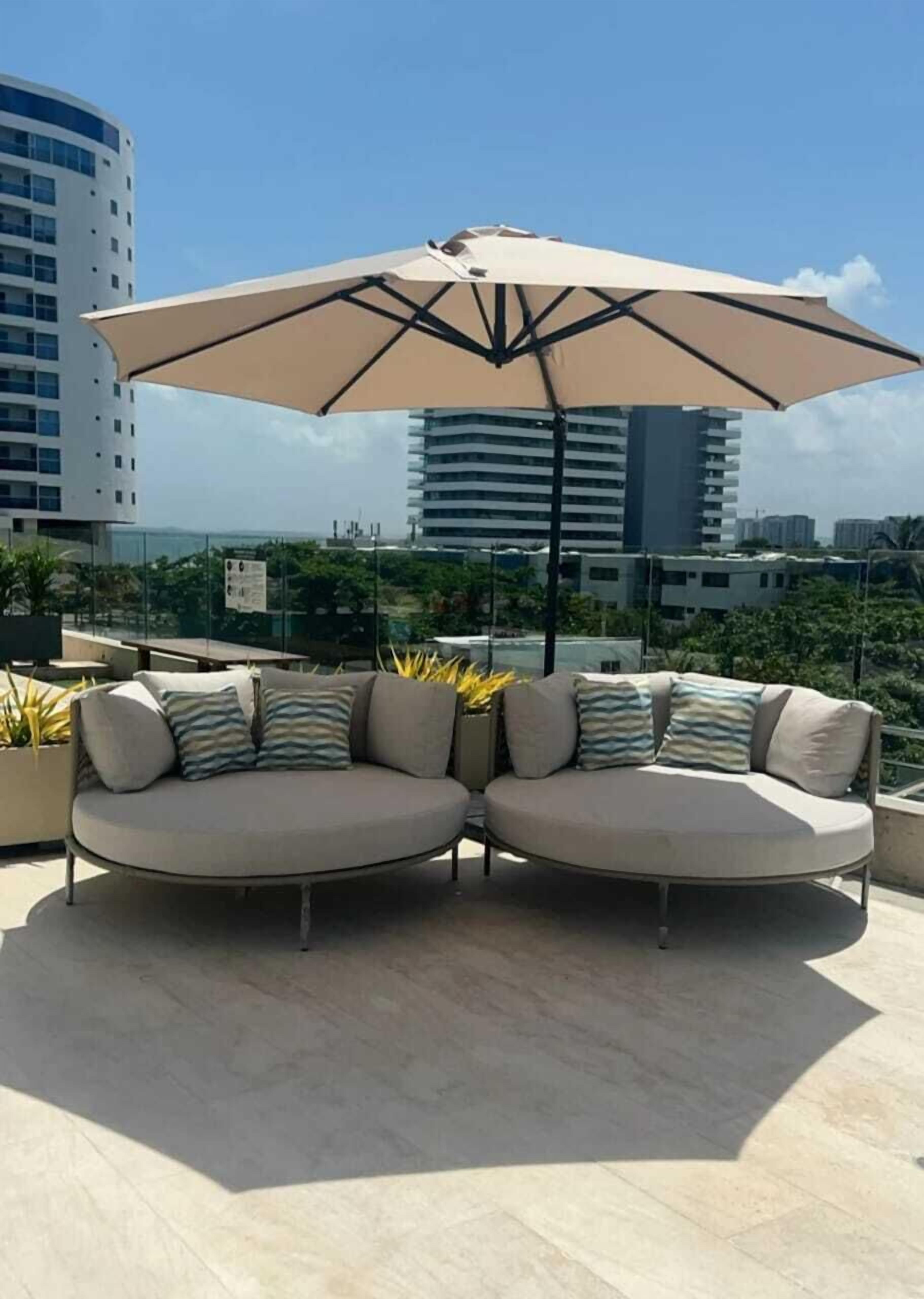 Un espace de détente aménagé sur le toit, avec un canapé beige et un grand parasol, entouré de plantes en pot et offrant une vue sur le paysage urbain.
