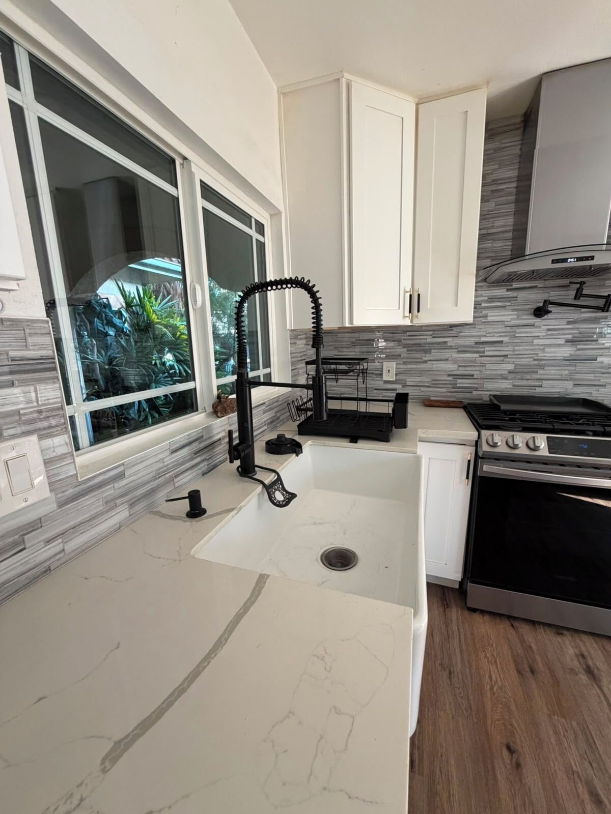 A modern kitchen with a large window, white cabinetry, a stainless steel stove, and a marble backsplash.