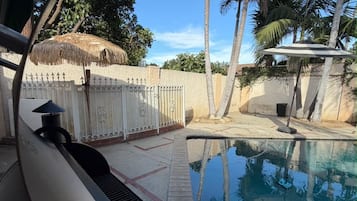 Ein Schwimmbecken mit weißem Zaun, Palmen und einer Struktur mit einem Reetdach.