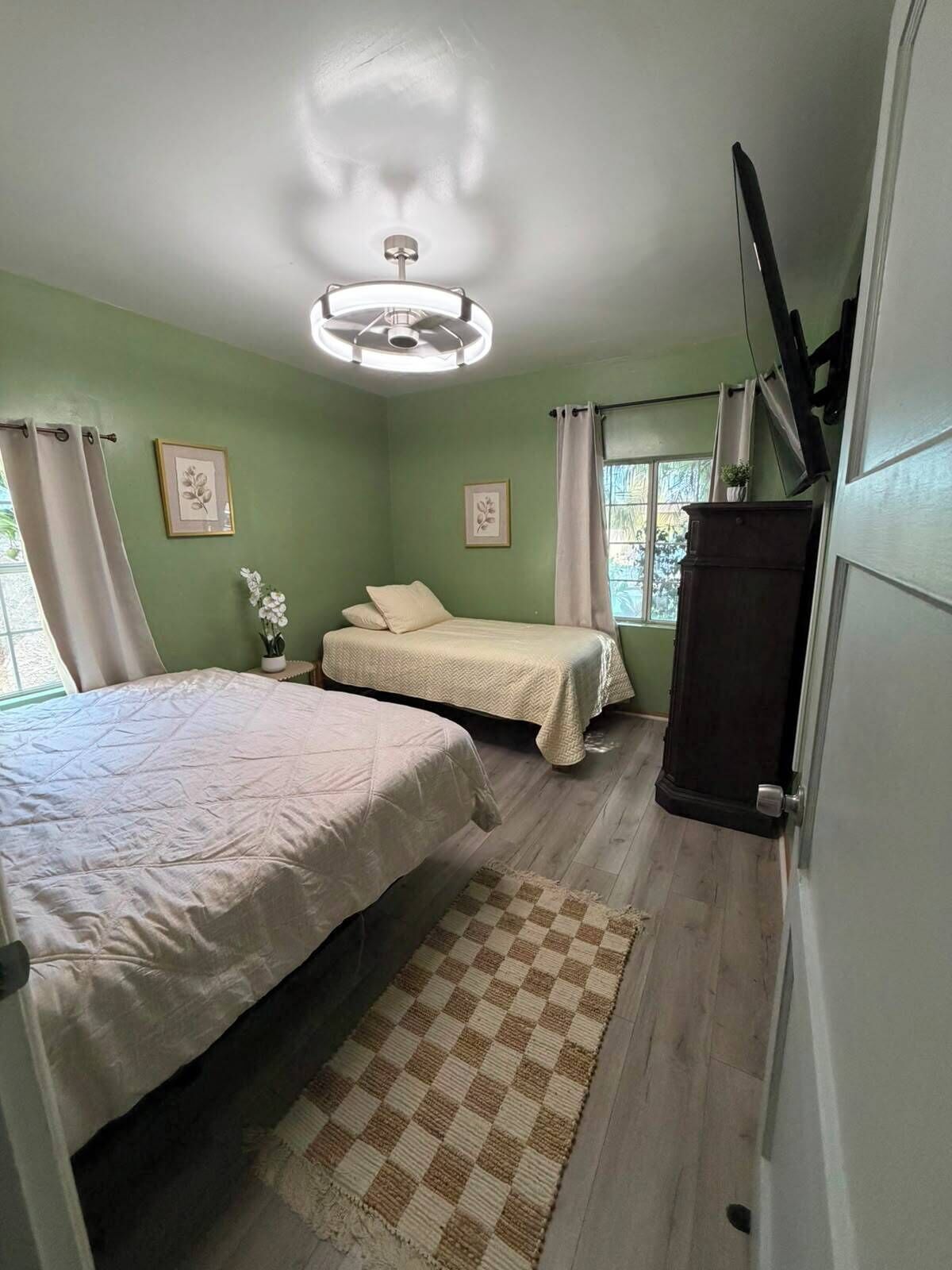 A bedroom with two beds, a ceiling fan, a window with curtains, and a checkered rug.