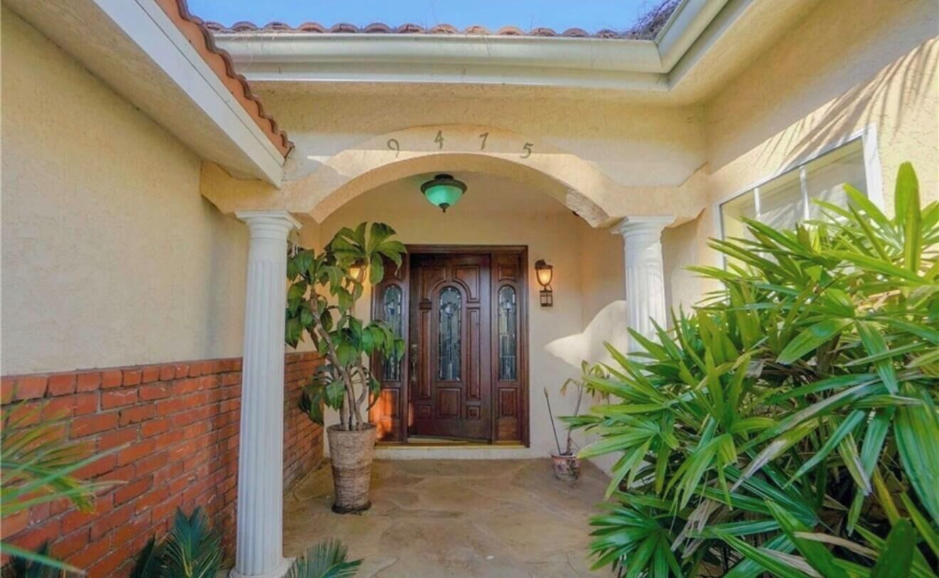 A building entrance with a number '9475' above the door, a brick wall to the left, and potted plants around.