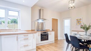A modern kitchen with white cabinetry, a stainless steel range hood, and an oven. There is a dining area with a table set for two and dark chairs. - Seascape Retreat - 3 Bedroom Flat - Porthcawl (Porthcawl)