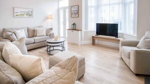 A living room with a sofa, armchair, and ottoman, a coffee table, a TV on a stand, and artwork on the walls. - Seascape Retreat - 3 Bedroom Flat - Porthcawl (Porthcawl)
