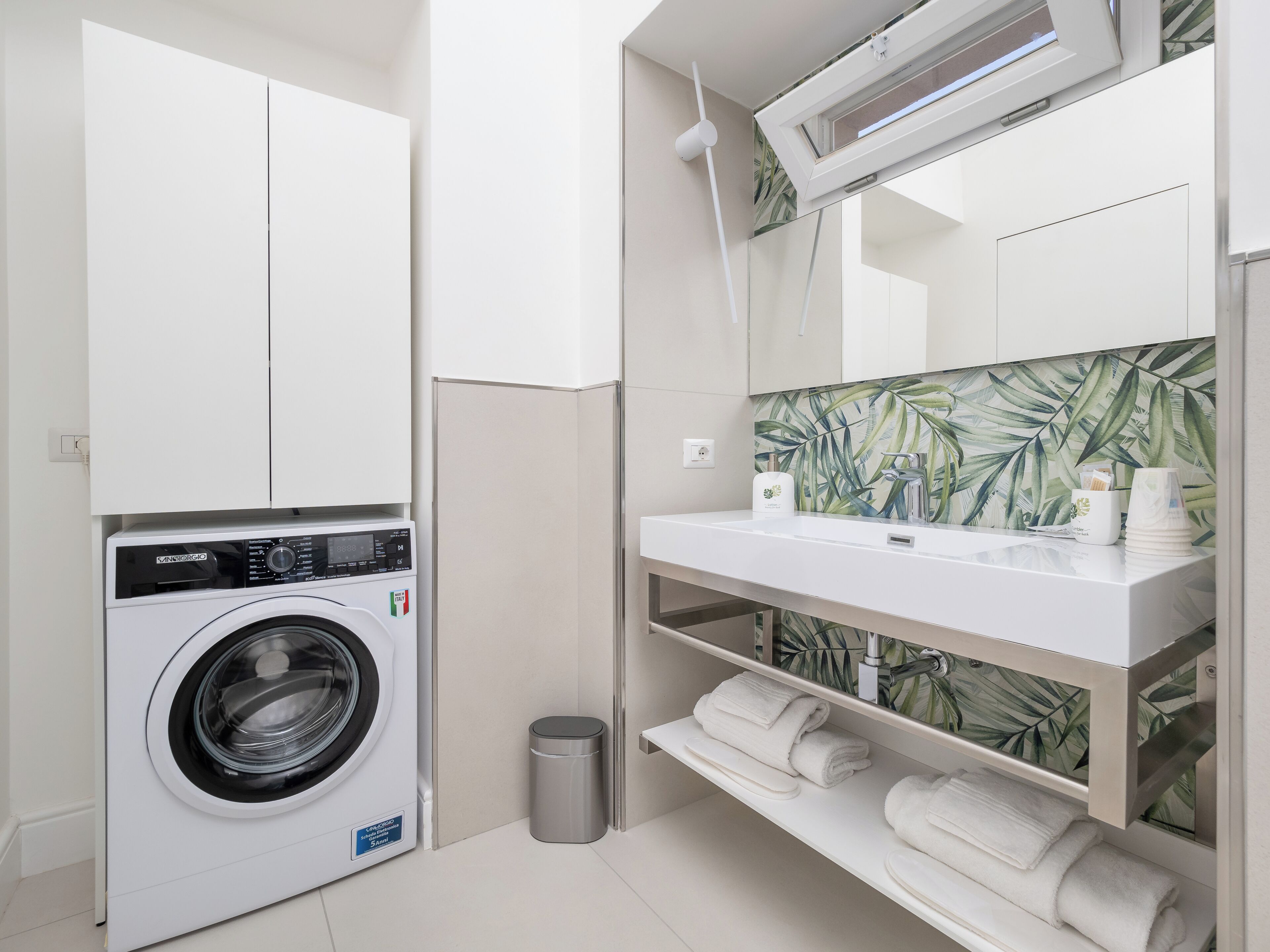 A modern laundry room with a front-loading washing machine, a small trash bin, and a white sink with a mirror above it.