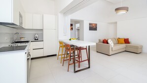 Una cocina moderna con armarios blancos, un comedor con mesa blanca y taburetes, y una sala de estar con sofá color beige y cojines coloridos.