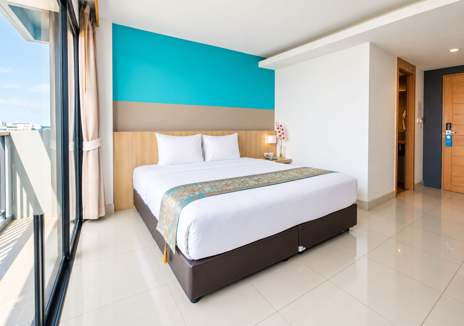 A hotel room with a large bed, a nightstand, a lamp, and a view of the city.