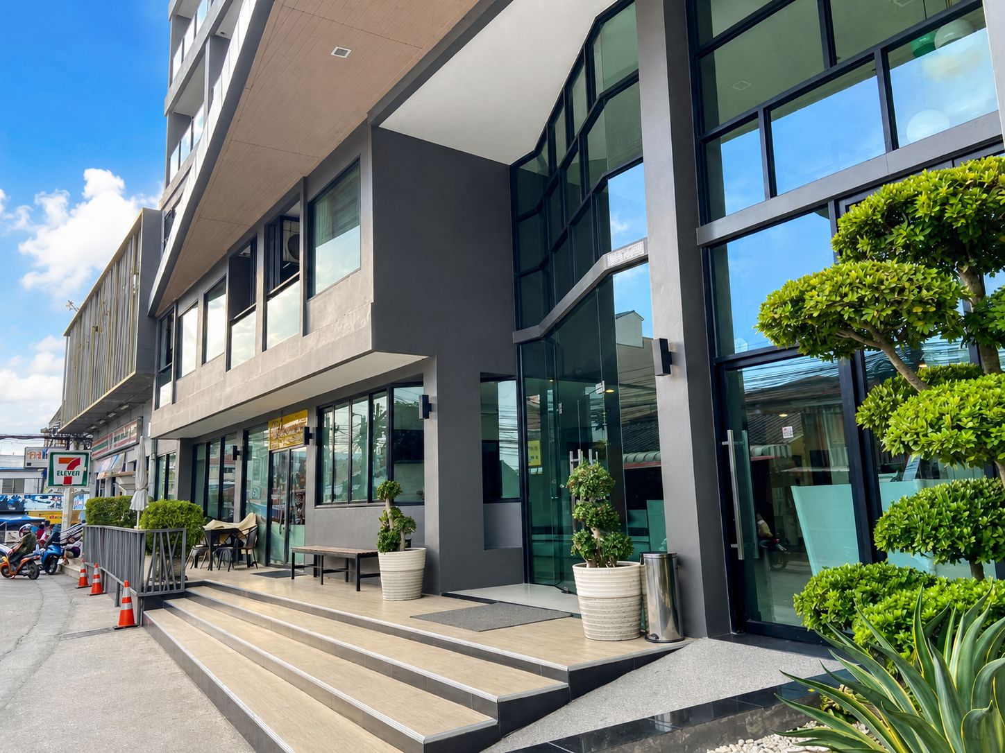 Un edificio moderno con techo plano, ventanas amplias y un área de entrada pavimentada con escalones y plantas en macetas.