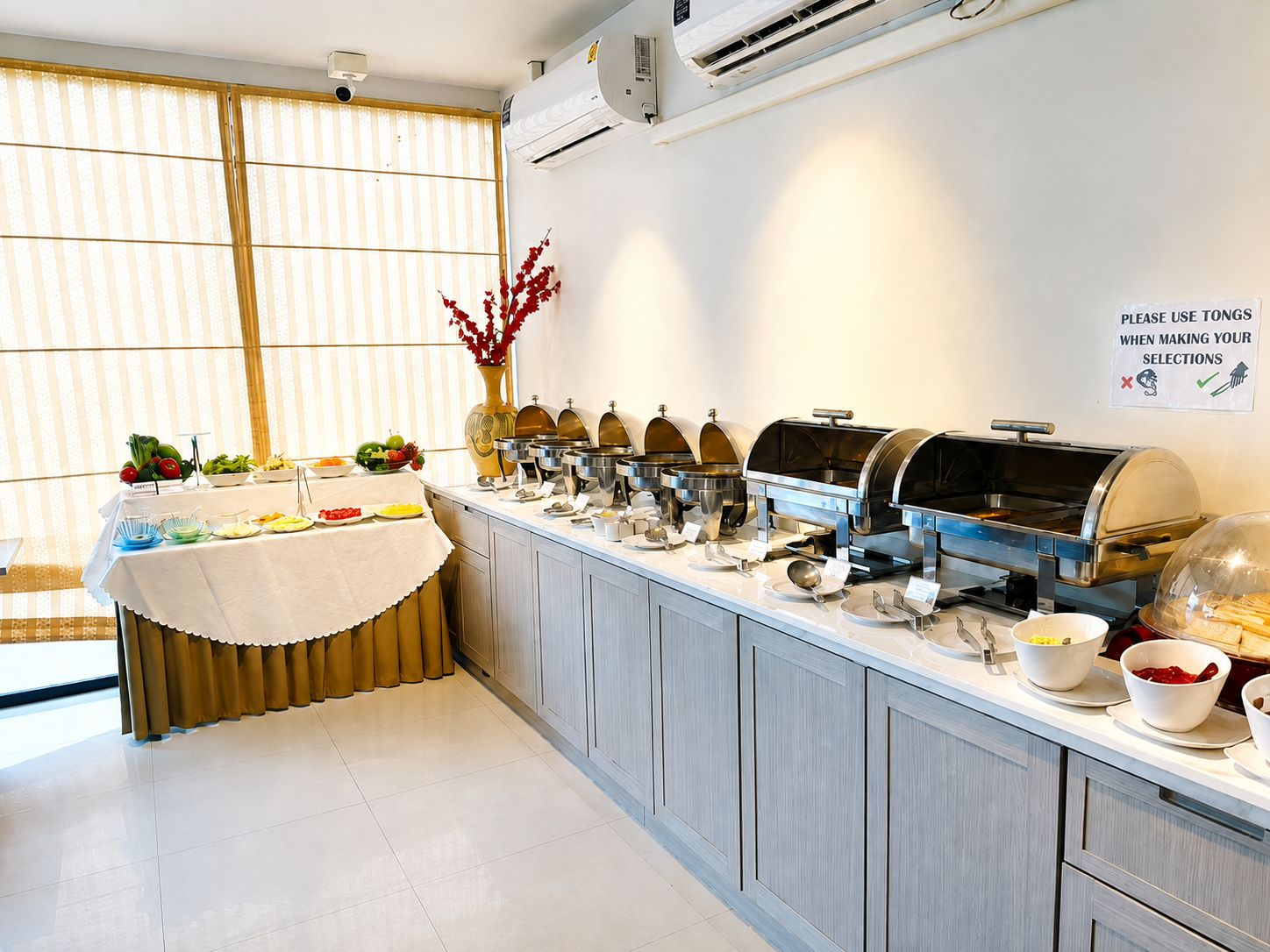 A buffet setup with multiple food stations, a sign instructing to use tongs, and a decorative vase with red flowers.