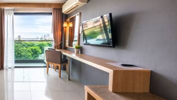 A modern hotel room with a large window, a flat-screen TV mounted on the wall, a wooden desk, and a chair.