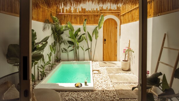 Área privativa da piscina com porta de madeira, cercada por plantas tropicais e um telhado de palha.