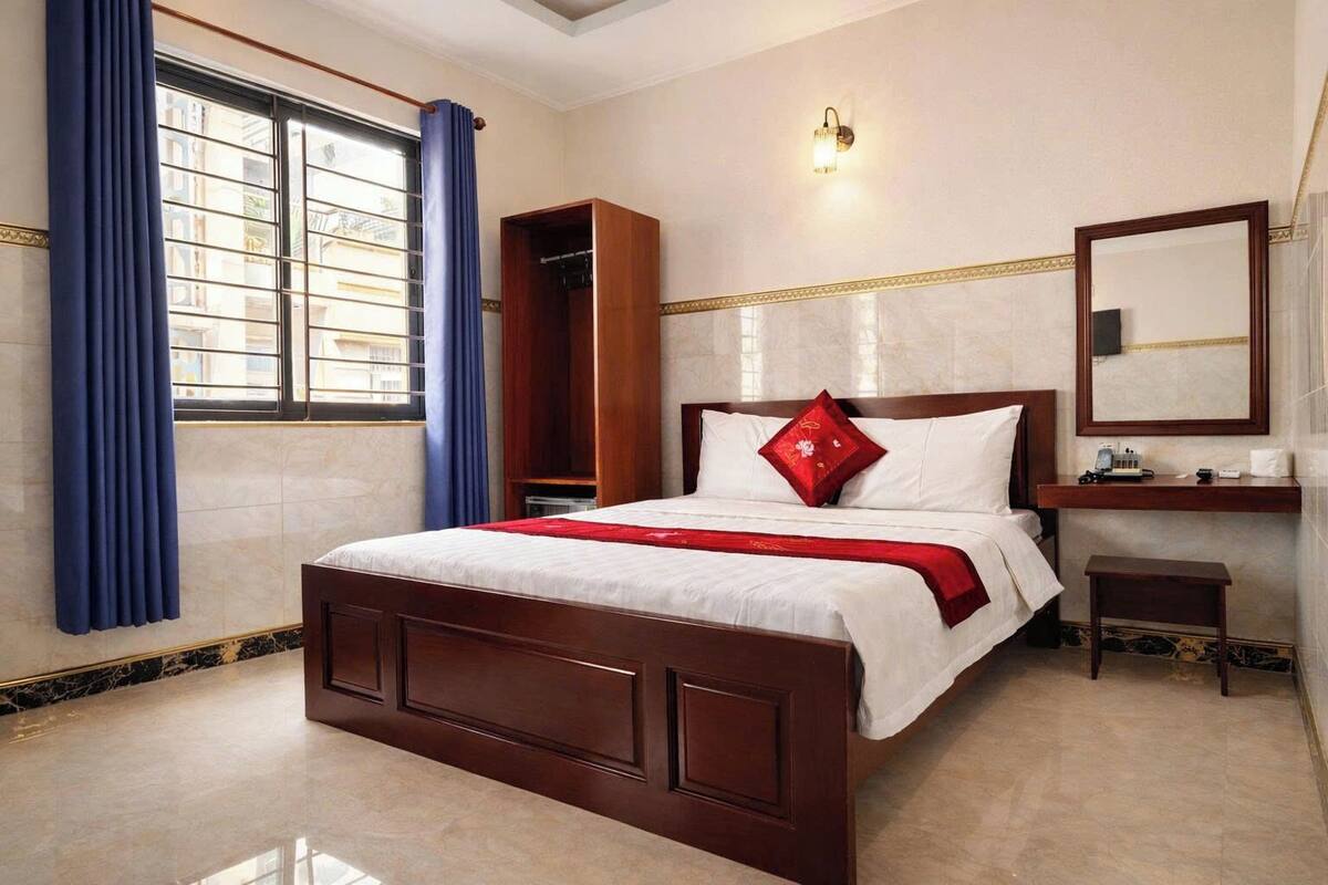 A hotel room with a large bed, a wooden wardrobe, a bedside table, a mirror, and a window with curtains.