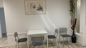 Un coin repas avec une table blanche et quatre chaises, une œuvre d’art encadrée au-dessus de la table et une plante en pot près de la fenêtre.