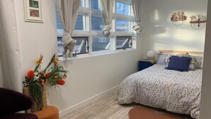 Une chambre à coucher comprenant un lit, une chaise, une petite table, une fenêtre avec des rideaux et un élément décoratif fixé au mur.