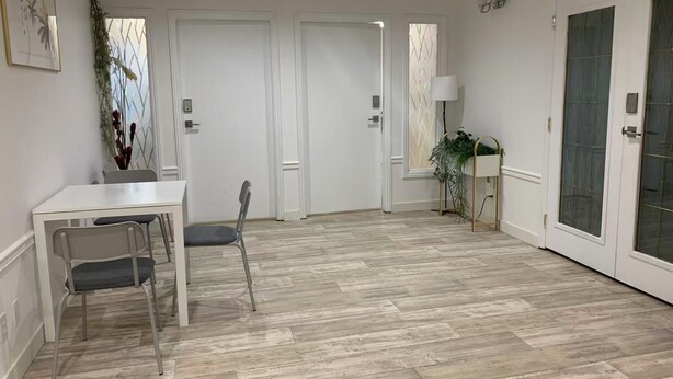 Une pièce spacieuse avec un plancher en bois, une table blanche avec deux chaises, une plante en pot et une porte dont les panneaux sont en verre dépoli.