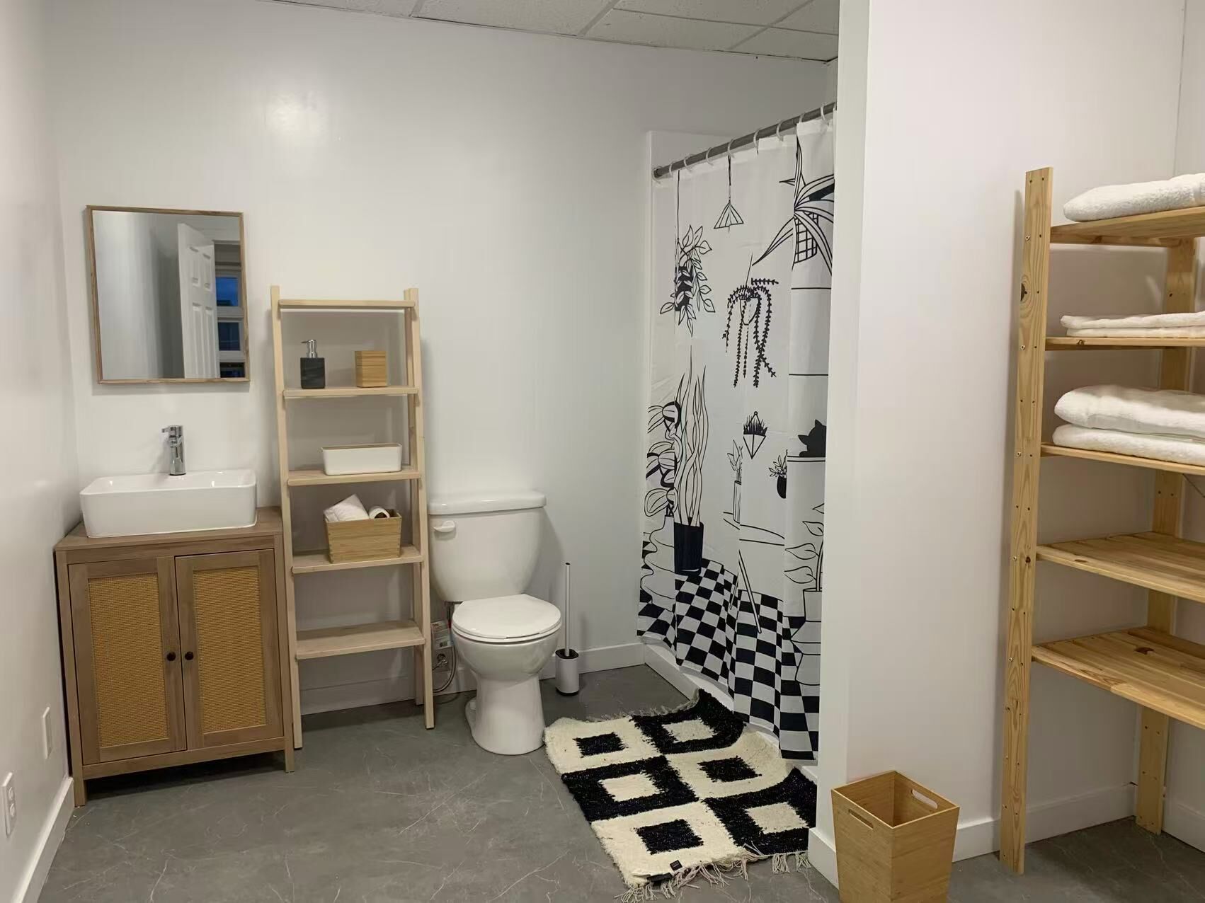 A bathroom with a white sink, wooden vanity, shelves, a toilet, a black and white checkered rug, and a shower curtain with plant designs.
