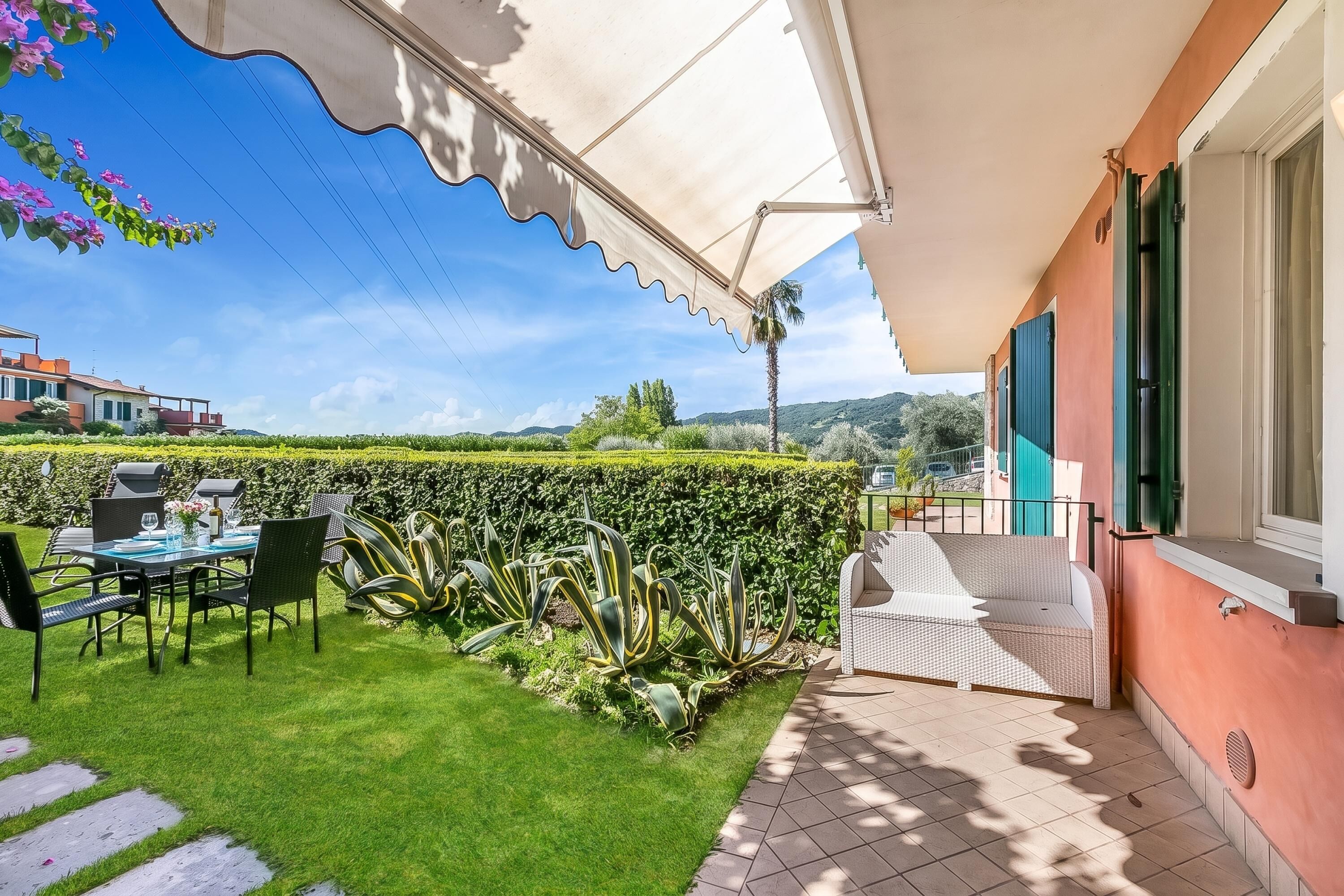 Un patio con una mesa y sillas, un sofá de mimbre blanco, y vistas a un jardín y colinas.