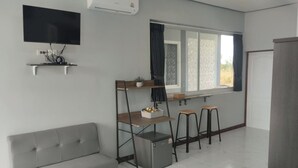 A compact living space with a grey sofa, a small kitchenette, a TV mounted on the wall, and a window with curtains.