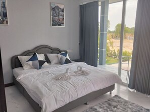 A spacious bedroom with a large bed, a view of the outdoors, and a cityscape painting on the wall. - The Village (Prakhon Chai)
