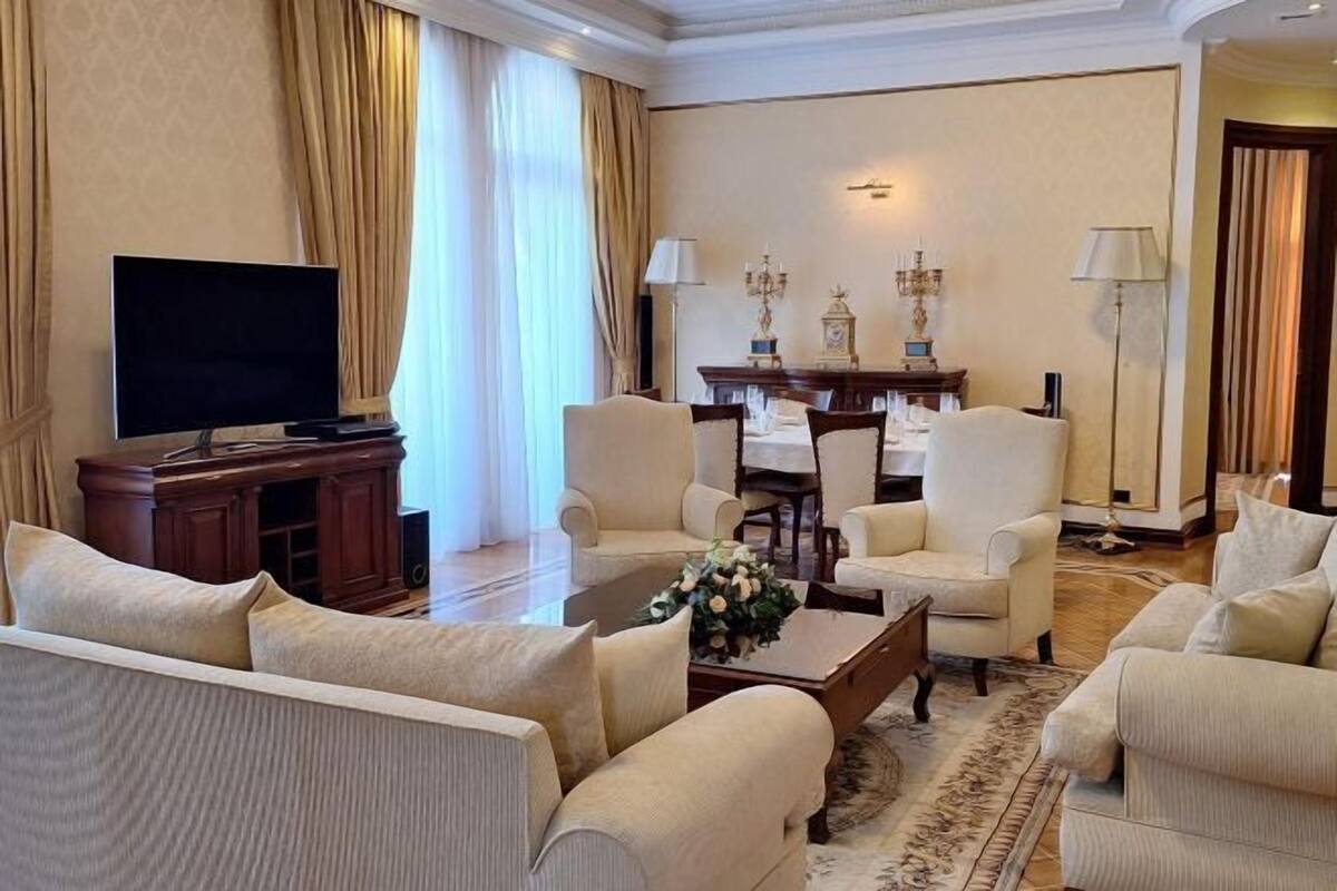 A living room with a flat-screen TV, a coffee table with flowers, and a dining area with a chandelier.