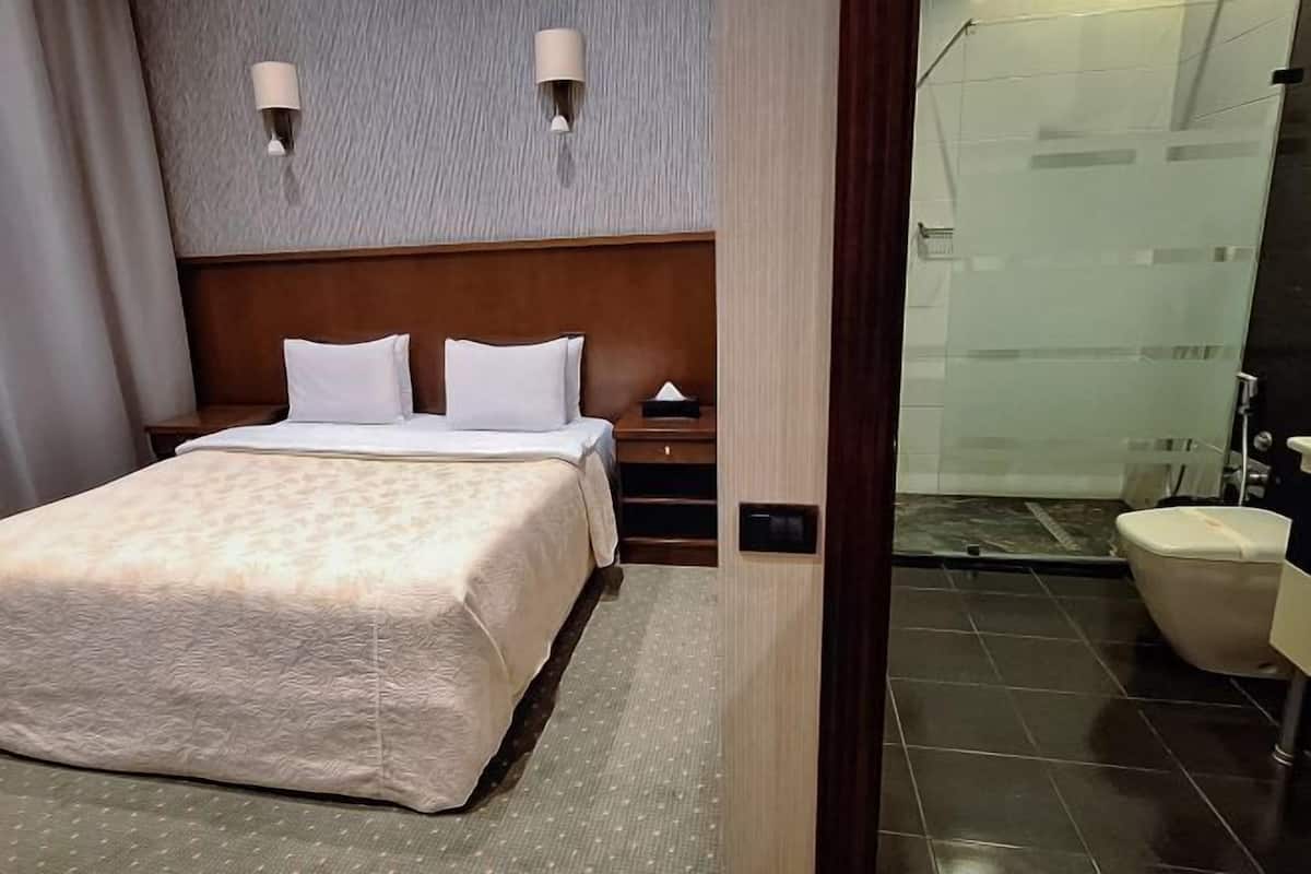 A hotel room with a bed, bedside tables, a nightstand, a wall-mounted lamp, and a bathroom with a toilet and shower.