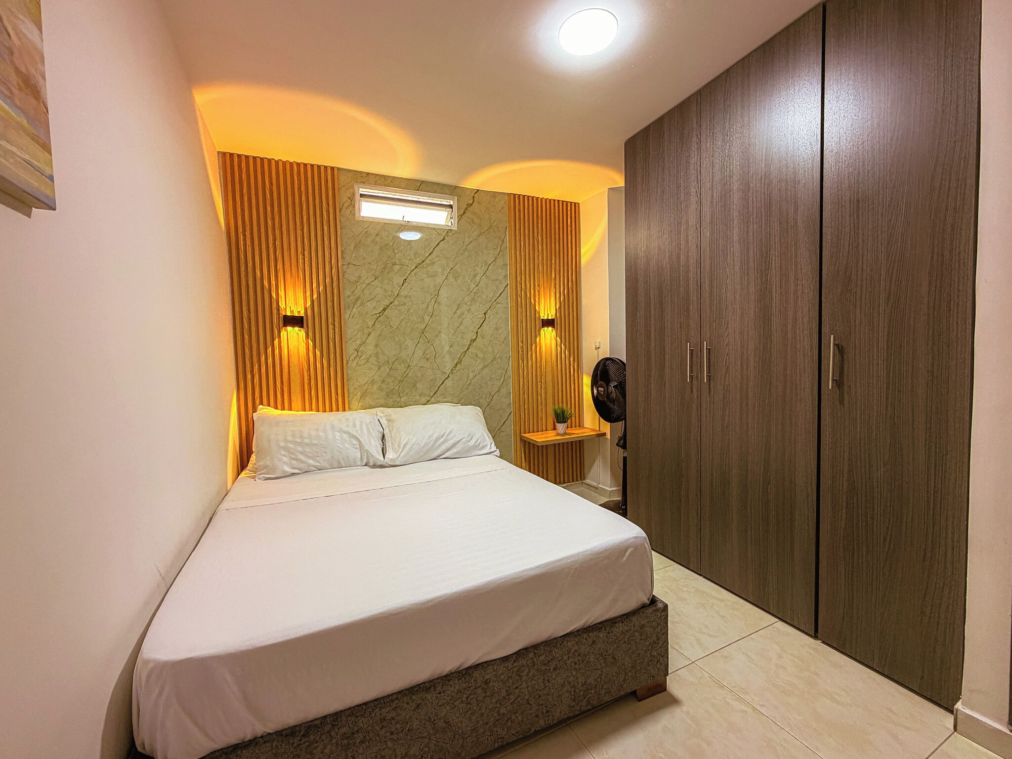A bedroom with a bed, wooden wardrobe, and a wall-mounted light fixture.