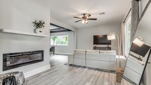 Una sala de estar moderna con chimenea, ventilador de techo, televisor de pantalla plana y un ventanal grande.