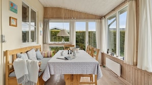 Un coin repas accueillant avec une table et des bancs en bois, de grandes fenêtres avec des rideaux et une vue sur la verdure environnante.